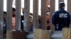 FILE - Migrants look through a border wall as a U.S. Customs and Border Protection officer, right, stands on the other side, seen from San Diego, Nov. 25, 2018.